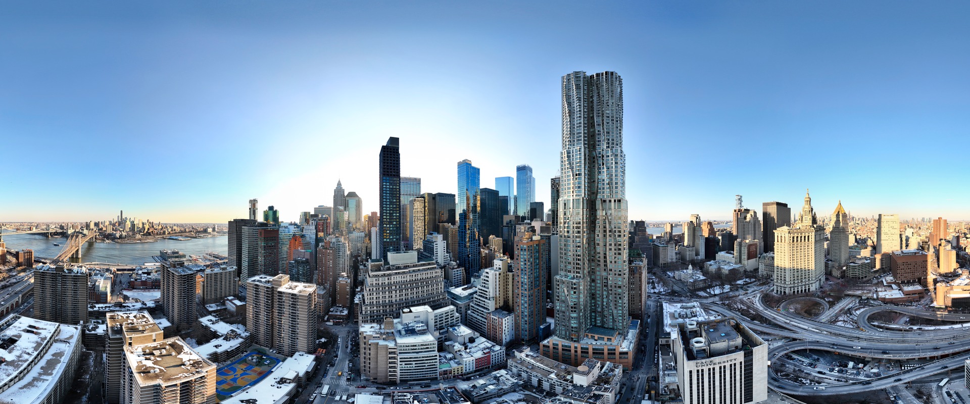 Panoramic winter view of Lower Manhattan skyline, featuring iconic towers, bridges, and the East River.